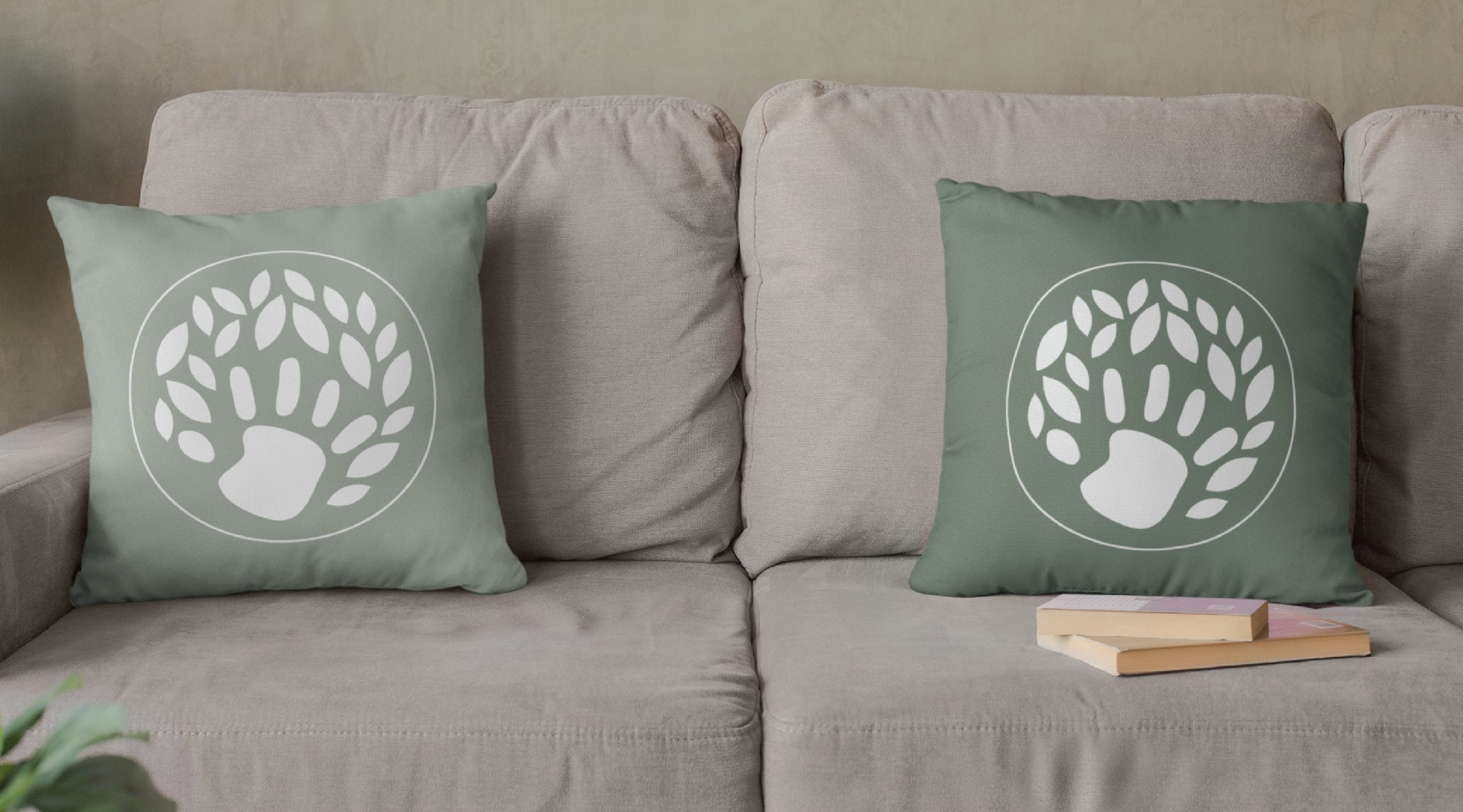 Two green throw pillows with white bear paw and leaf designs—reminiscent of a playful logo design for a children’s therapist in Virginia—sit on a beige couch. A couple of books are stacked on the right side of the couch cushion.