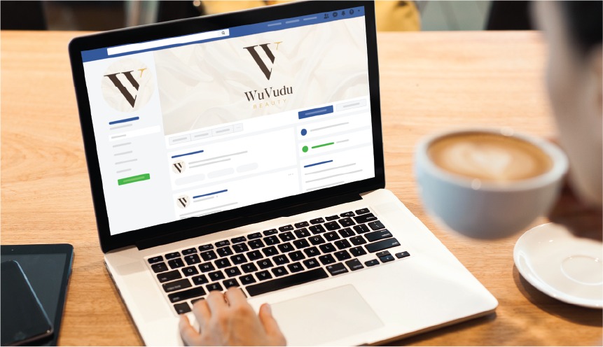 A person uses a laptop displaying a Facebook business page for WuVudu Beauty, showcasing expert social media design, while holding a cup of coffee at a wooden table with a phone and notebook nearby in Indiana.