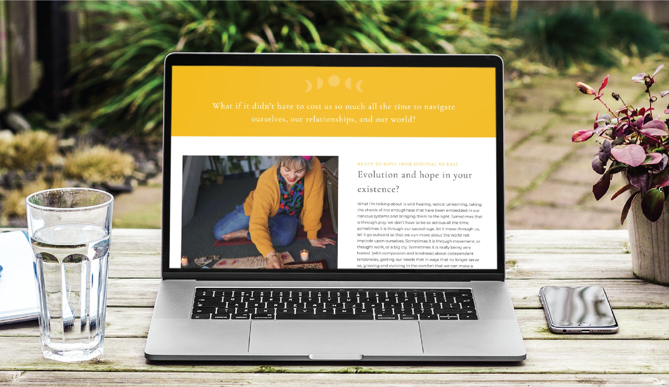 A laptop on an outdoor table displays a website for a Spiritual Therapist with a yellow header and a photo of a woman sitting on a rug. A glass of water, a smartphone, and a notebook are beside the laptop, creating an inviting Colorado work setting.