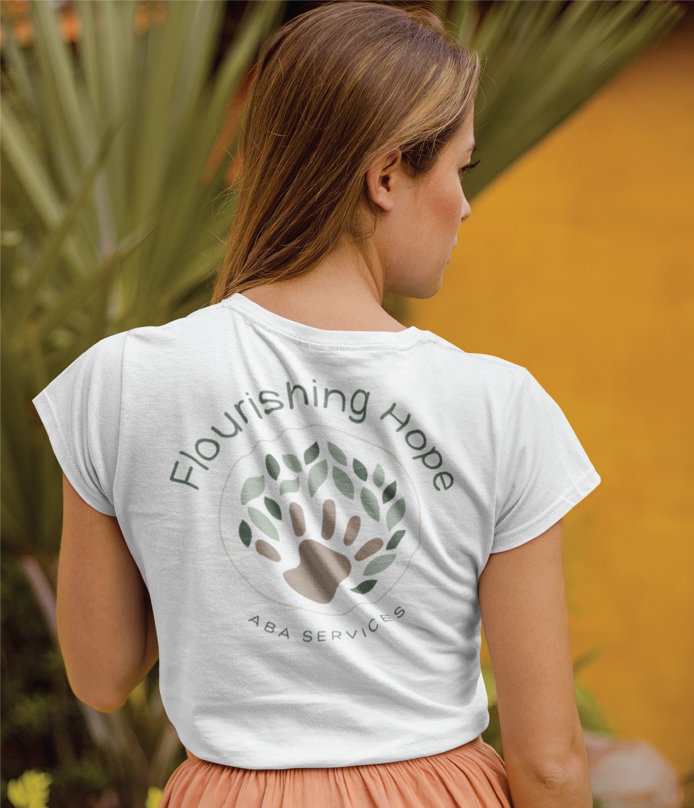 A woman with light brown hair faces away, wearing a white shirt that says Flourishing Hope ABA Services with a logo design featuring a hand surrounded by leafy branches. Green plants and a yellow wall are in the background, hinting at the welcoming atmosphere often found in a children’s therapist office in Virginia.