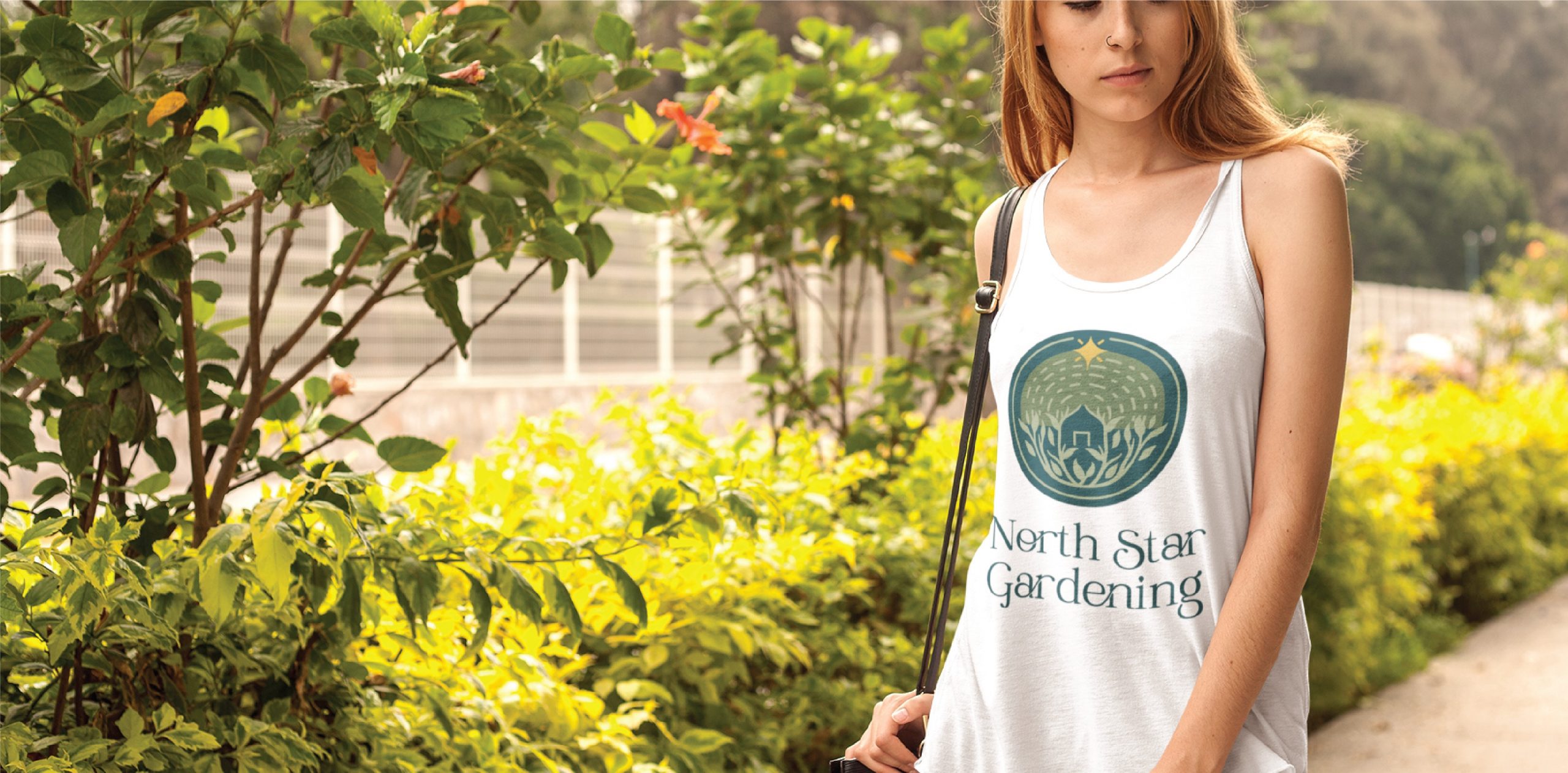 A woman stands in a California garden wearing a white tank top with a green North Star Gardening logo illustration, surrounded by lush plants and flowers.