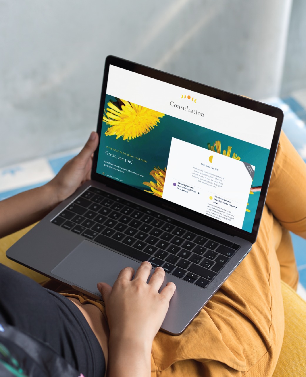 A person sits cross-legged holding a laptop on their lap, viewing a beautifully crafted Website Design for a Colorado Spiritual Therapist, featuring vibrant yellow dandelion flowers and a Consultation section on the screen.
