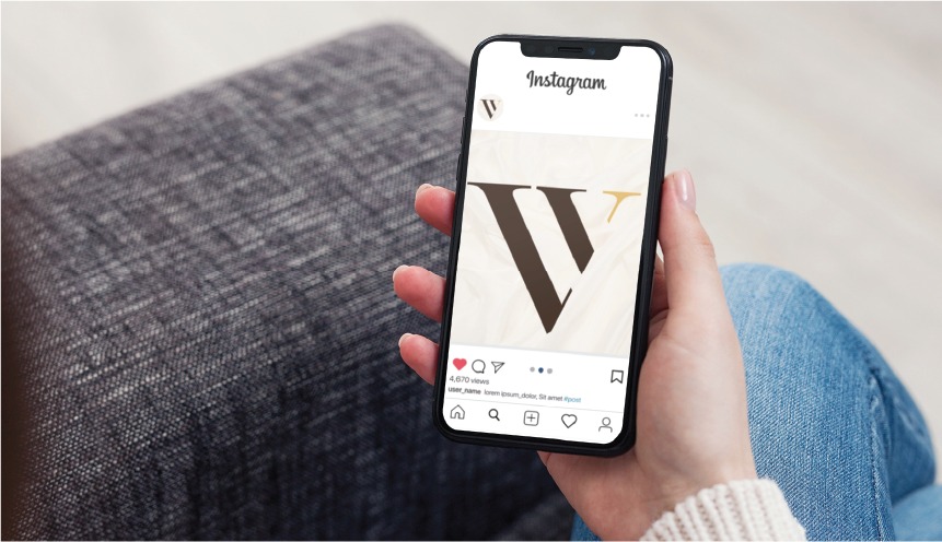 A person holds a smartphone featuring an Instagram post with a large stylized W on a beige background, showcasing thoughtful Social Media Design. The phone rests on their lap above a gray sofa.