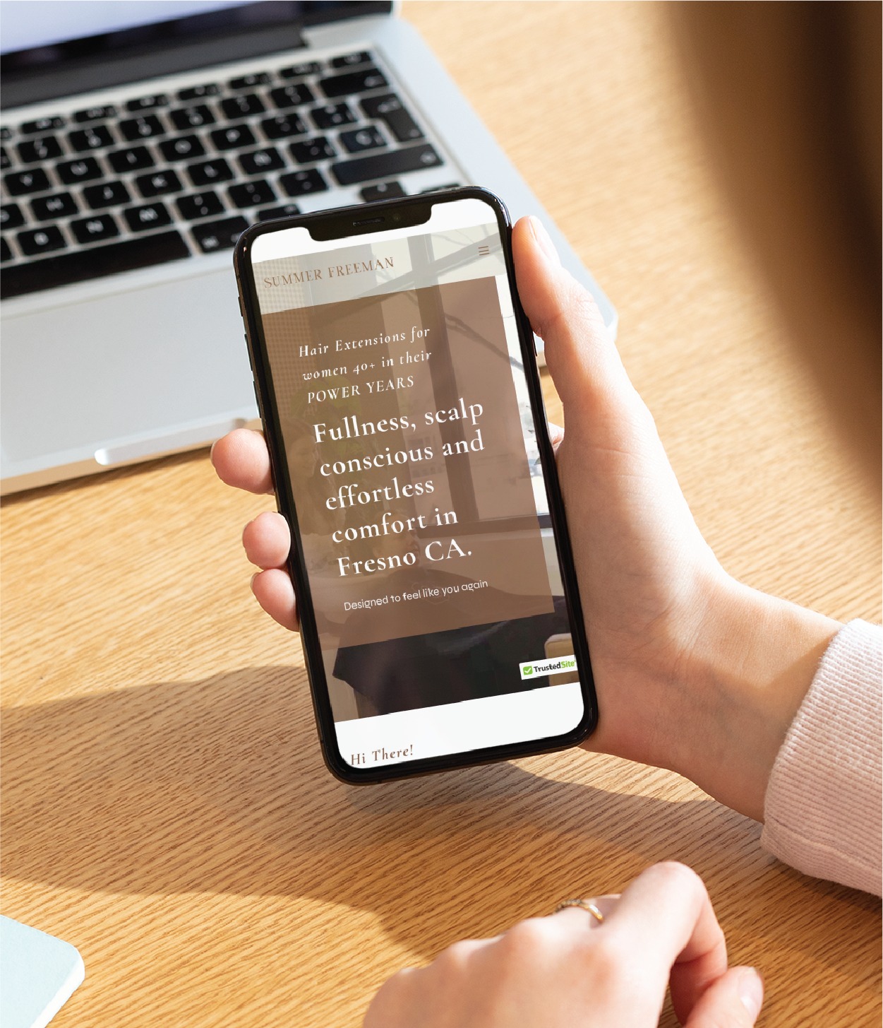 A person holds a smartphone displaying a beautifully designed website about hair extensions for women, emphasizing fullness and comfort in Fresno, California. The phone—possibly belonging to a local hairdresser—is held above a wooden desk.