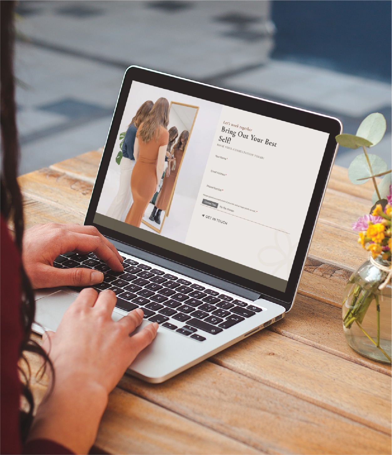 A person types on a laptop at a wooden table, viewing a website design featuring a photo of a woman in a tan dress looking in a mirror. A vase with flowers is nearby, creating a relaxed California vibe.