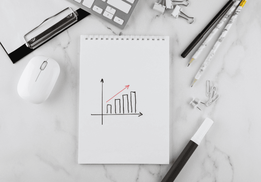 A notepad with a bar graph showing growth, surrounded by a mouse, clipboard, pens, binder clips, and paperclips on a white desk—ideal for organizing your marketing tracking efforts and visually monitoring link tracking progress.