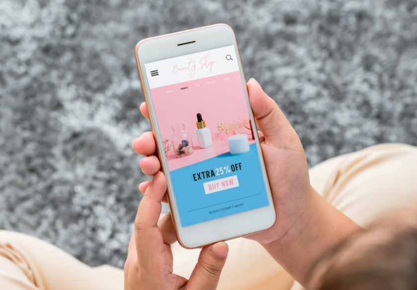 A person holds a smartphone displaying a beauty shop landing page with a pink and blue theme, showing skincare products and a banner for EXTRA 25% OFF and a BUY NOW button—perfect for discovering small business brands.