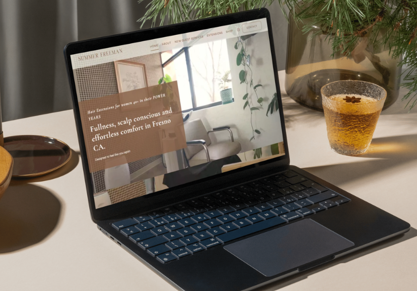 A black laptop on a beige desk displays a salon website you can trust. Next to it are a drink in a textured glass, a wooden coaster, and a pine branch in a glass vase. Sunlight casts soft shadows on the scene, creating an inviting atmosphere where you can trust your website to shine.