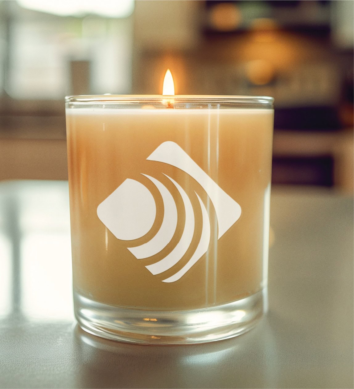 A lit beige candle in a clear glass holder sits on a smooth surface, featuring a white geometric logo with curved lines—a subtle nod to thoughtful branding design. The background shows a softly blurred indoor setting, evoking the comforting atmosphere often found in modern US mental health services.