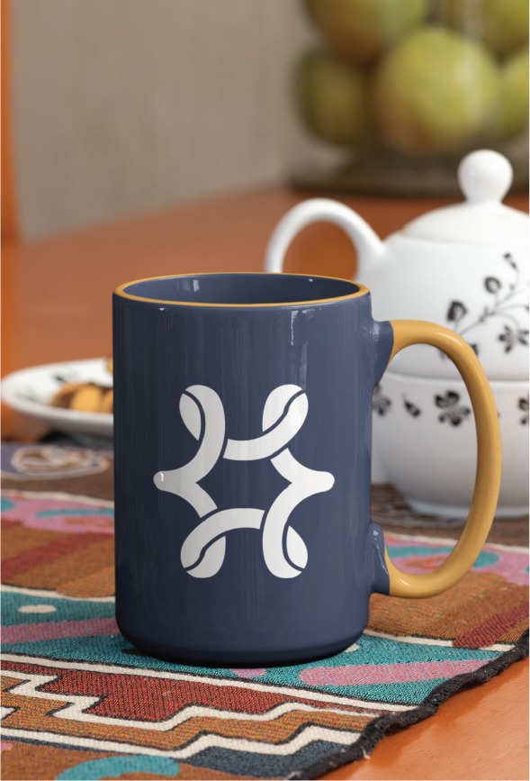 A navy blue mug with a yellow handle features a modern white abstract design inspired by the vibrant spirit of New Orleans. The mug sits on a colorful patterned cloth beside a white teapot, with a blurred bowl of fruit in the background.