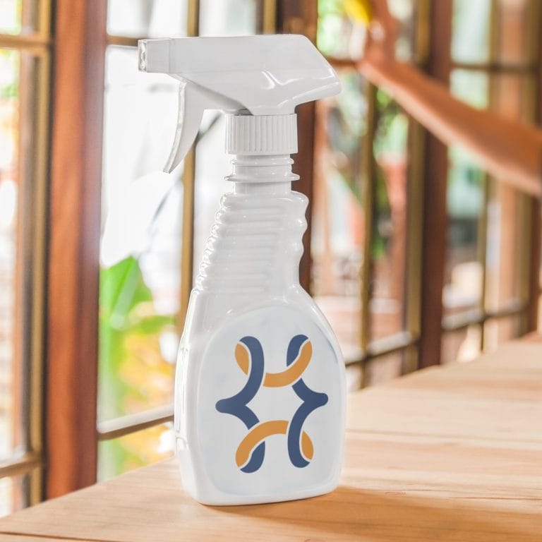 A white spray bottle with a blue and orange abstract design sits on a wooden table in a sunlit room, evoking the vibrant spirit of New Orleans, with a blurred arm and windows in the background.