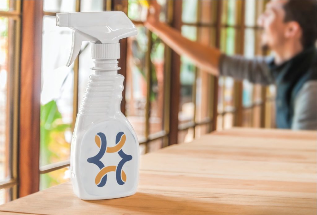 A white spray bottle with a blue and orange logo sits on a wooden table, while a person cleans large windows in the blurred background—capturing a bright day in New Orleans, just steps away from Tulane University.