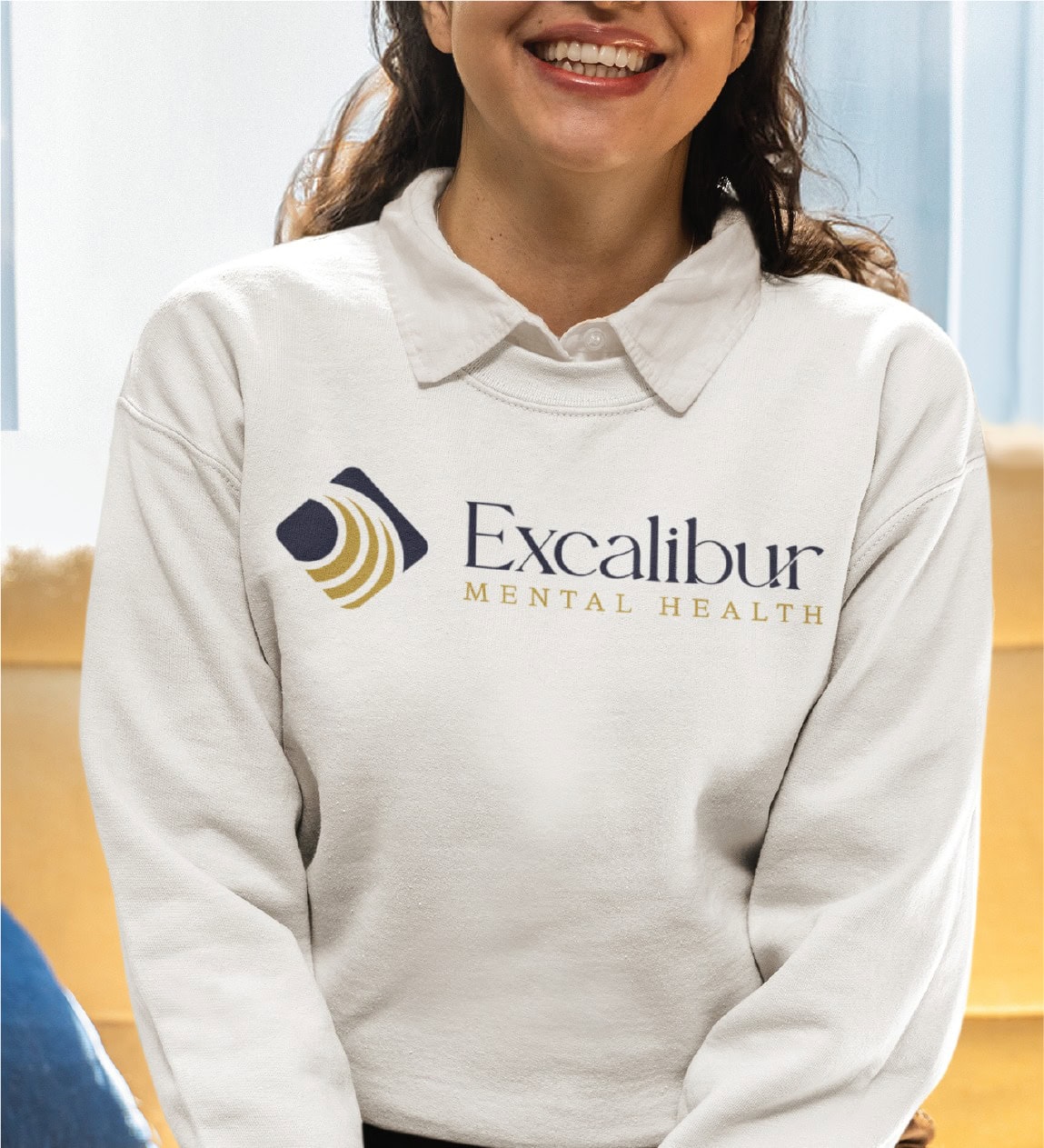 A smiling person wearing a white collared sweatshirt with the Excalibur Mental Health logo and text printed on the front, showcasing thoughtful branding design for mental health services in the US.