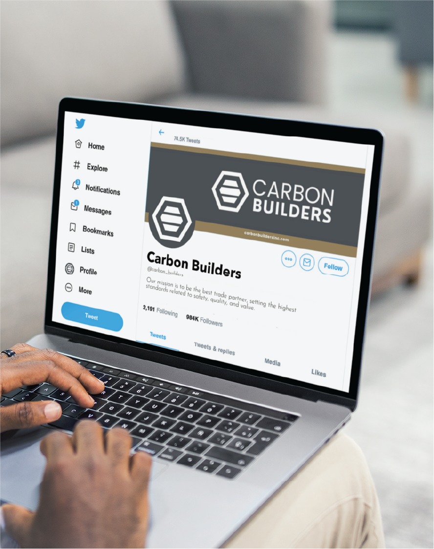A person using a laptop views the Twitter profile of Carbon Builders, a Full Service Contractor based in Michigan. The profile features a banner and logo with the company name, custom social media design elements, and icons for notifications, messages, and other Twitter sections.