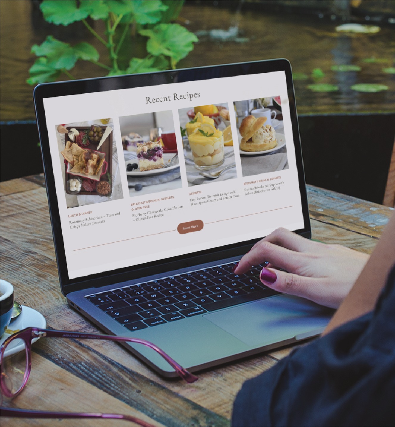 A person uses a laptop outdoors, viewing a food website featuring three recent recipe posts with images and descriptions—perfect inspiration for any home cook. Pink glasses rest beside the laptop on a wooden table, capturing the relaxed vibe of California living and the appeal of custom website design.