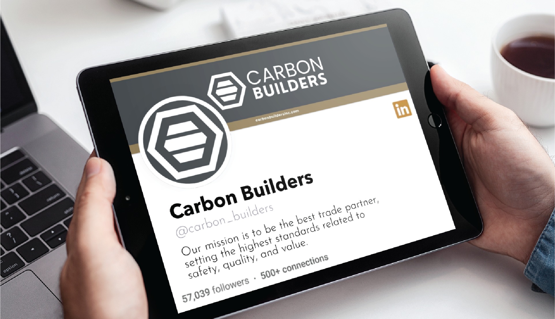 A person holds a tablet displaying the LinkedIn profile of Carbon Builders, a Full Service Contractor based in Michigan, showing their logo, tagline, and follower count above a laptop and coffee cup on a desk featuring Custom Social Media Design elements.