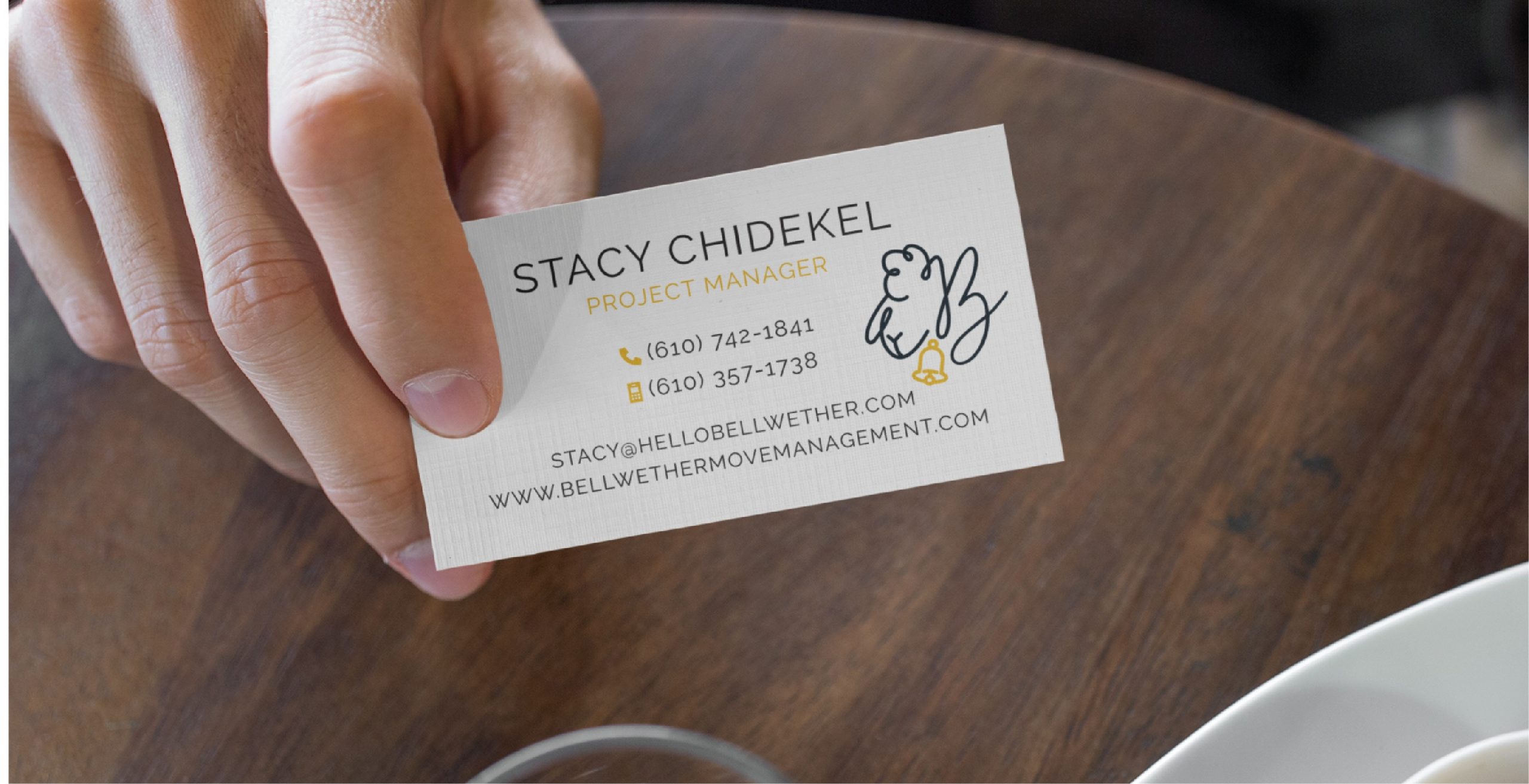 A hand holds a business card for Stacy Chidekel, Project Manager, featuring custom print design with phone numbers, email, website, and a stylized sheep logo on a wooden table near a white plate—a detail that highlights her expertise in move management across Pennsylvania.