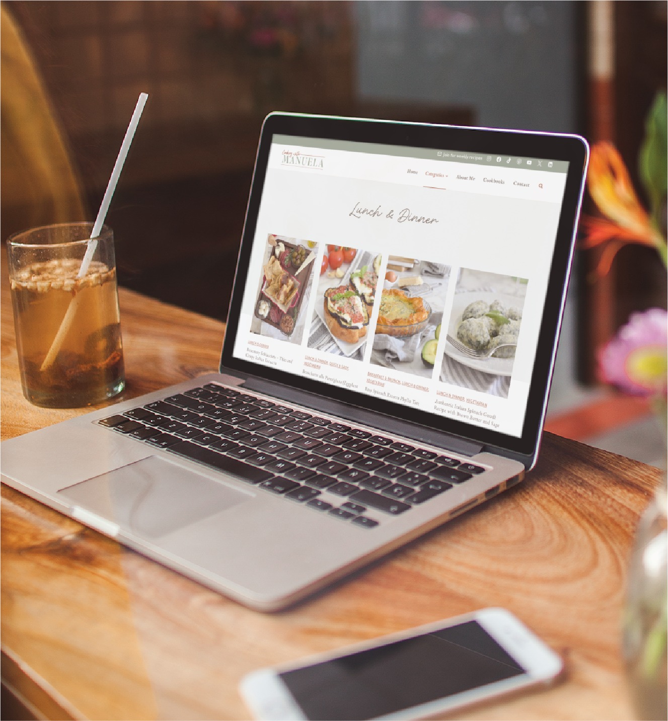 A laptop on a wooden table displays a food blog with lunch and dinner recipes, perfect for any passionate home cook. Next to it are a glass of iced drink with a straw and a smartphone. Sunlight streams in through the window, creating the relaxed vibe of a California afternoon.