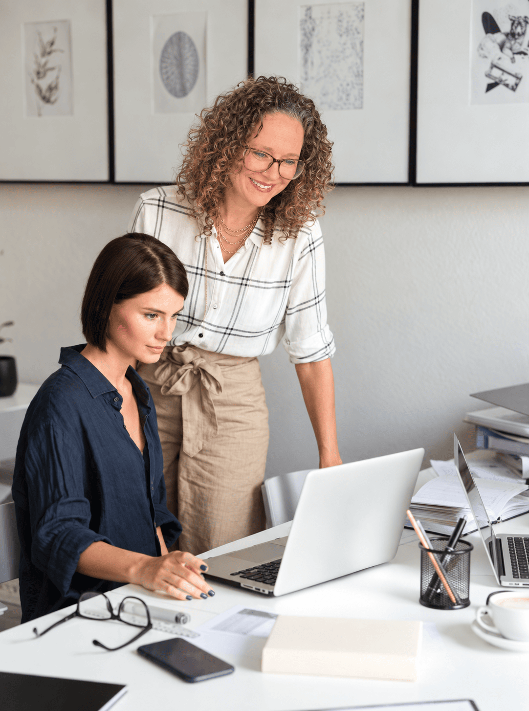 Two women working together at a desk with a laptop and papers, collaborating on website design. One types while the other smiles at the screen. The office features framed artwork in the background.