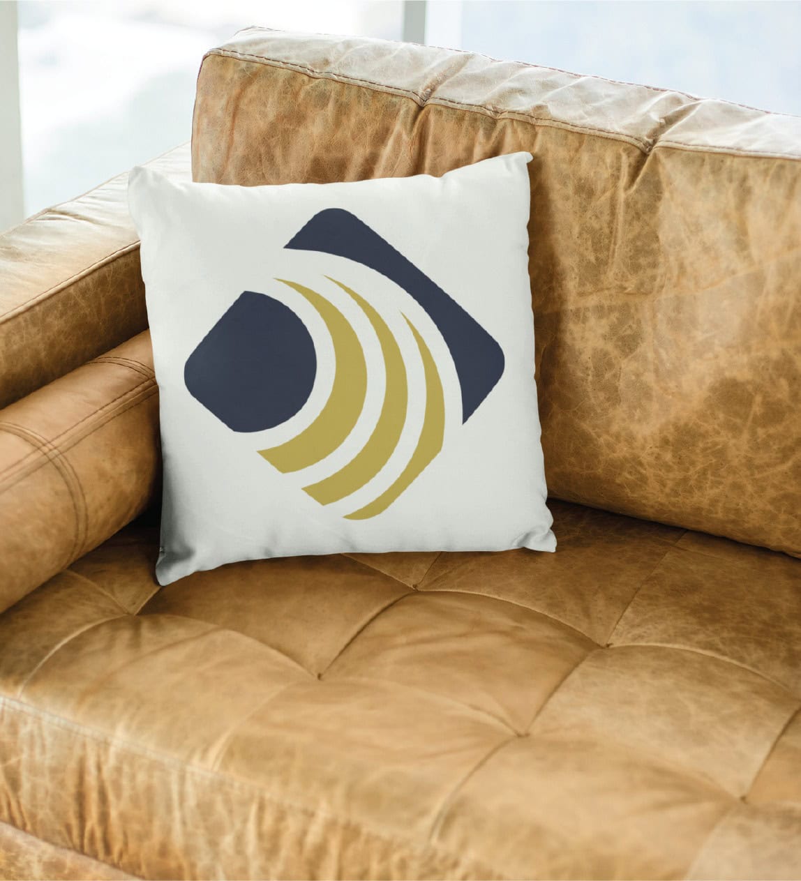 A white pillow with a modern abstract design in navy and gold rests on a light brown leather couch near a window, adding a touch of sophisticated branding design often seen in contemporary US interiors.