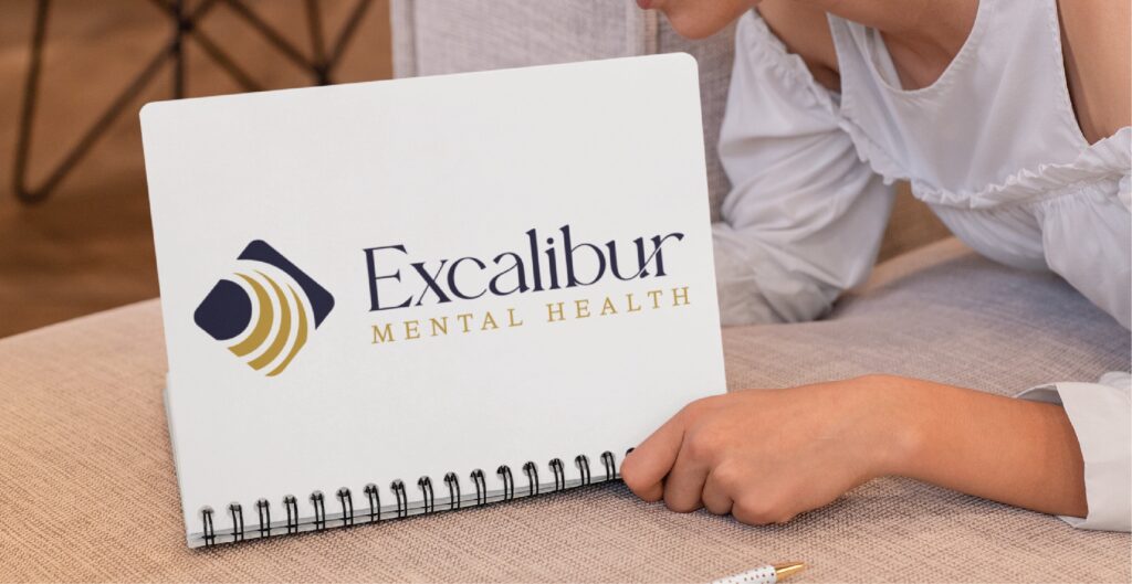 A person holds a spiral notebook displaying the Excalibur Mental Health logo, which features abstract gold and navy shapes next to the business name in navy and gold text. This striking branding design highlights Excalibur’s commitment to providing trusted mental health services in the US.