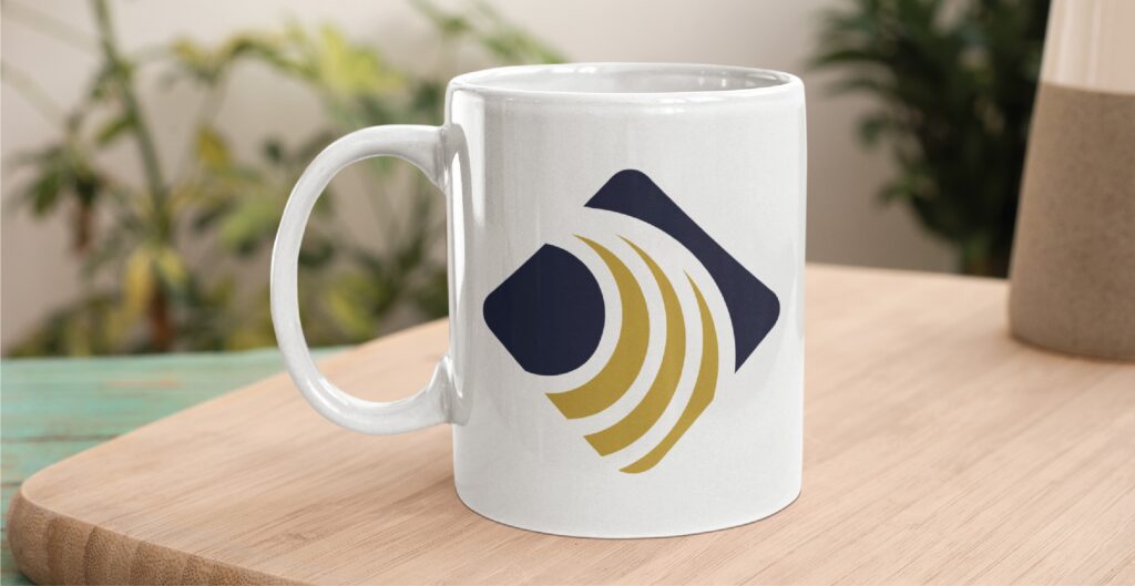 A white mug with a blue and gold abstract logo, inspired by modern branding design, sits on a wooden surface, with blurred green plants in the background.