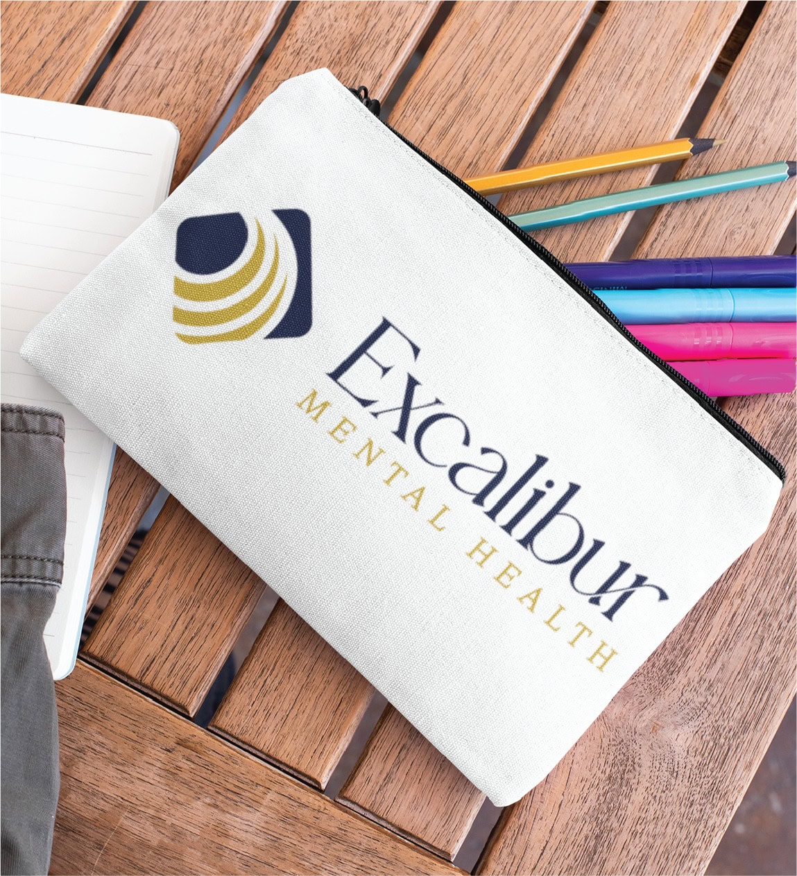 A white zippered pouch with the Excalibur Mental Health logo—showcasing thoughtful branding design—sits on a wooden table beside colorful pens, a pencil, a notebook, and part of a gray sleeve. This scene highlights the approachable side of US mental health services.