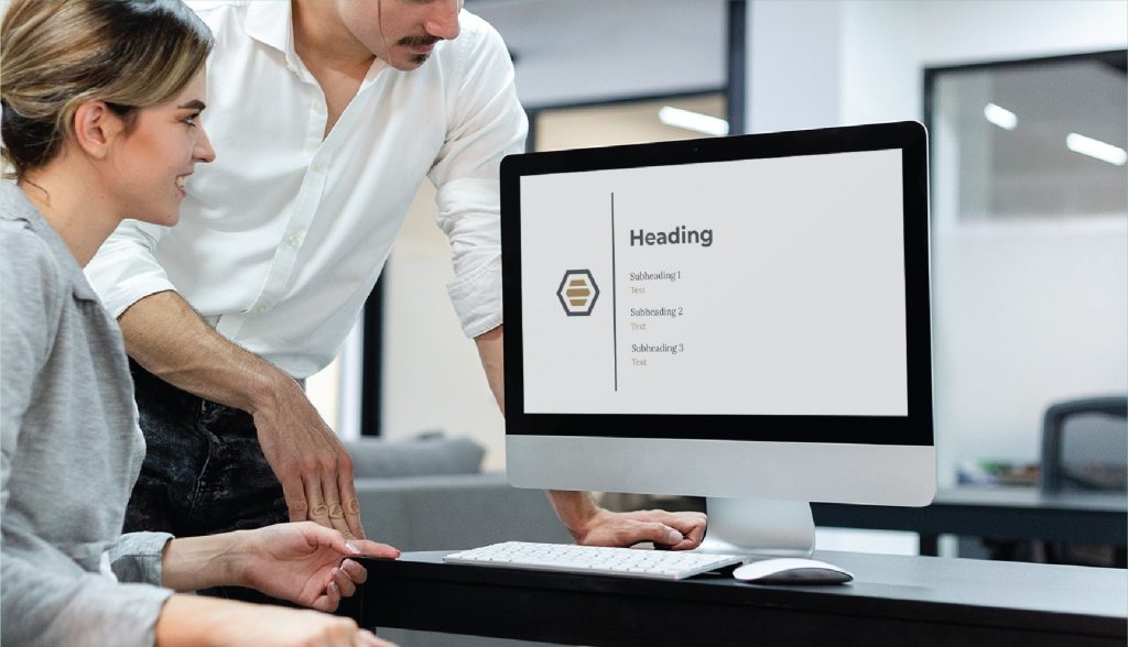 Two people in an office look at a computer screen displaying a simple presentation slide with the heading Heading and placeholder text for subheadings and text, discussing ideas for custom social media design for their Michigan full service contractor business.