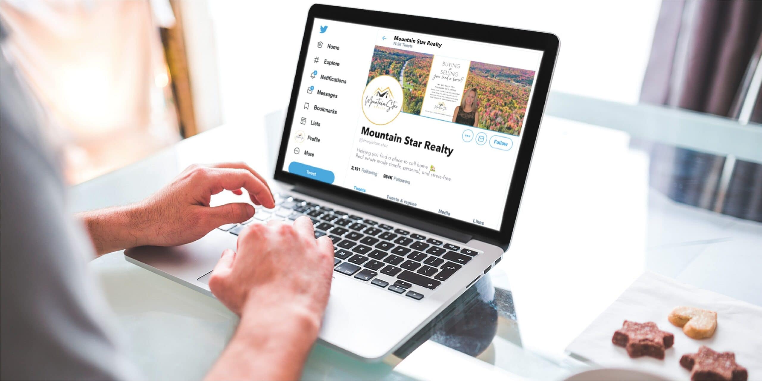 Person using a laptop with a Twitter profile for Mountain Star Realty open on the screen, showcasing their Custom Social Media Design; cookies and a drink are on the table nearby, creating a cozy workspace for this Michigan Contractor.