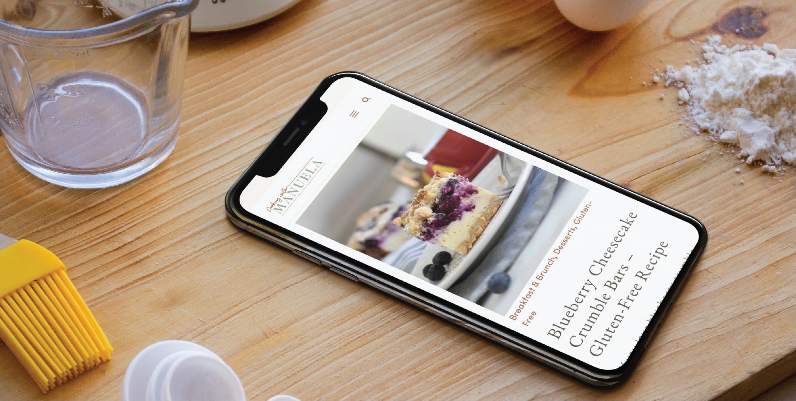 A smartphone on a wooden surface displays a blueberry cheesecake recipe, perfect for any aspiring California home cook. Baking tools, flour, and an egg are nearby, suggesting baking in progress.