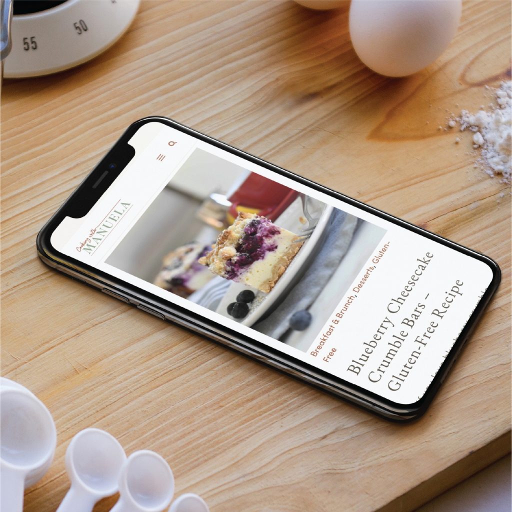 A smartphone displays a blueberry cheesecake crumble bar recipe on a custom website design catering to the home cook, sitting on a wooden surface next to a timer, measuring spoons, and spilled flour—perfectly capturing the relaxed vibe of California kitchens.