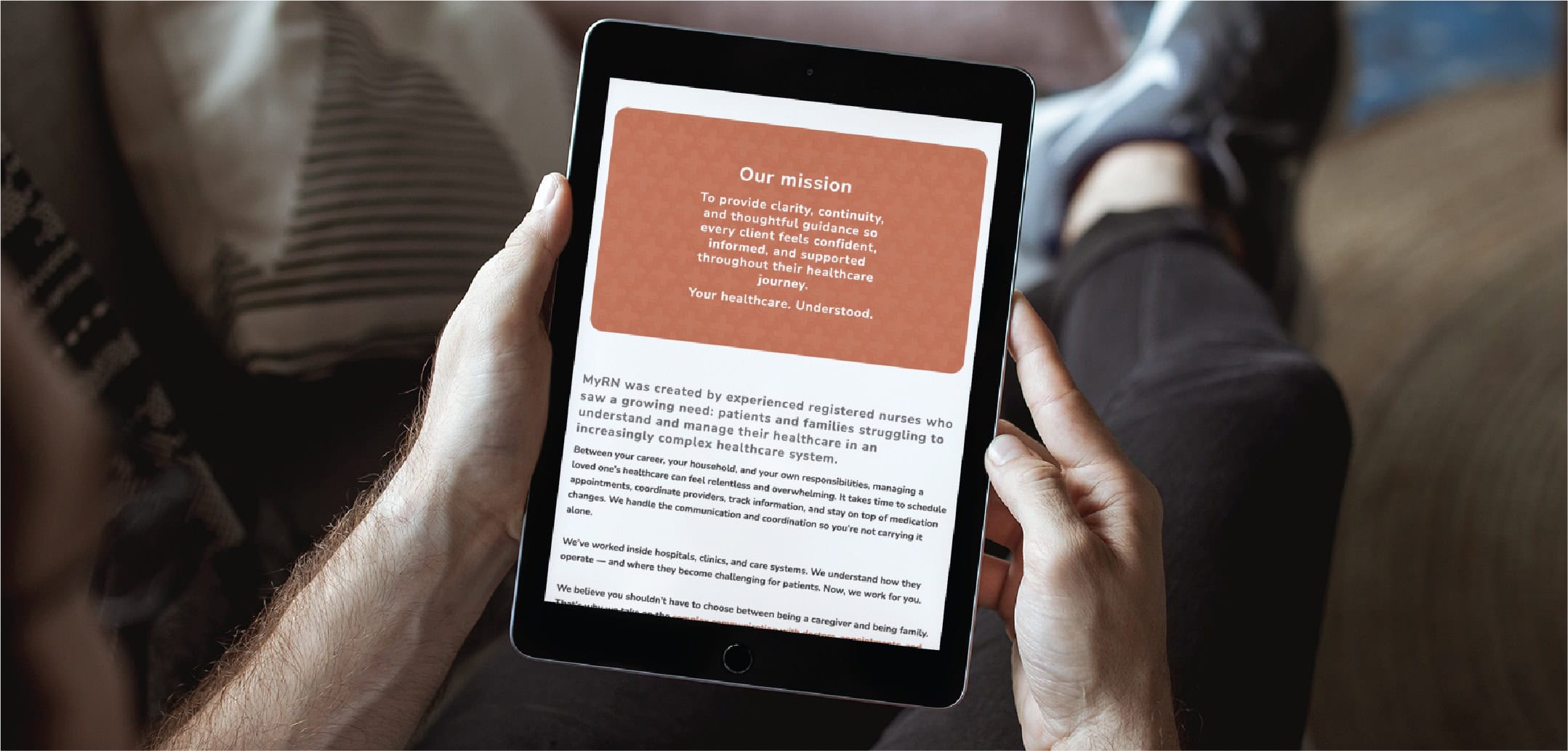 Person holding a tablet displaying a mission statement in white text on a red background, with additional black text below. The person is seated on a couch, and the softly blurred background suggests a comfortable Minnesota setting—ideal inspiration for Website Design or RN Services.
