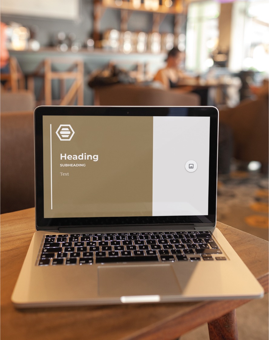 A laptop sits on a wooden table in a cafe, displaying a presentation slide with the word Heading and placeholder text. The blurred background shows people and seating, creating an inviting scene ideal for brainstorming custom social media design projects with your Michigan full service contractor.