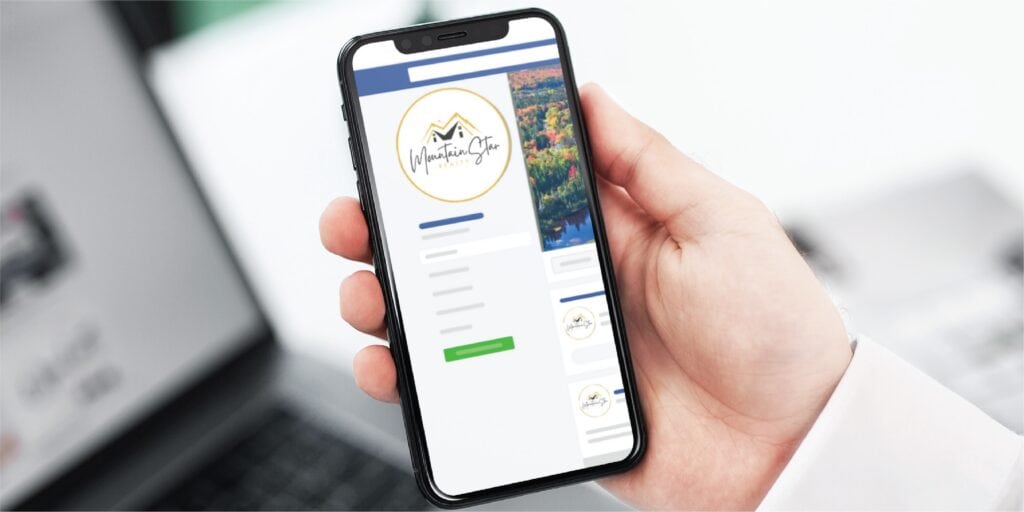 A person holds a smartphone displaying a social media profile with a circular logo, profile details, and a cover photo featuring a Michigan landscape. A blurred laptop and workspace are visible in the background, emphasizing the impact of Custom Social Media Design for any Full Service Contractor.