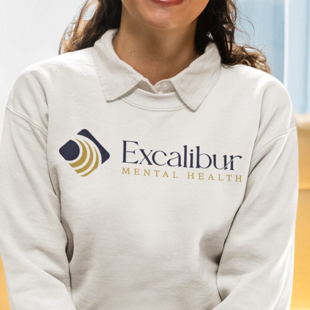 A woman wearing a white collared sweatshirt featuring the Excalibur Mental Health logo and text on the front, showcasing thoughtful branding design that represents US-based mental health services.