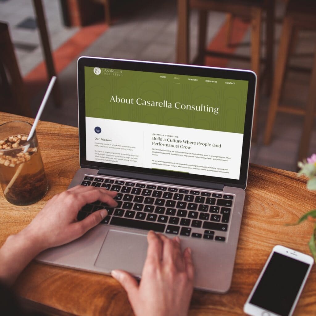 A person uses a laptop displaying the Casarella Consulting website design on a wooden table next to a glass of iced tea and a smartphone.