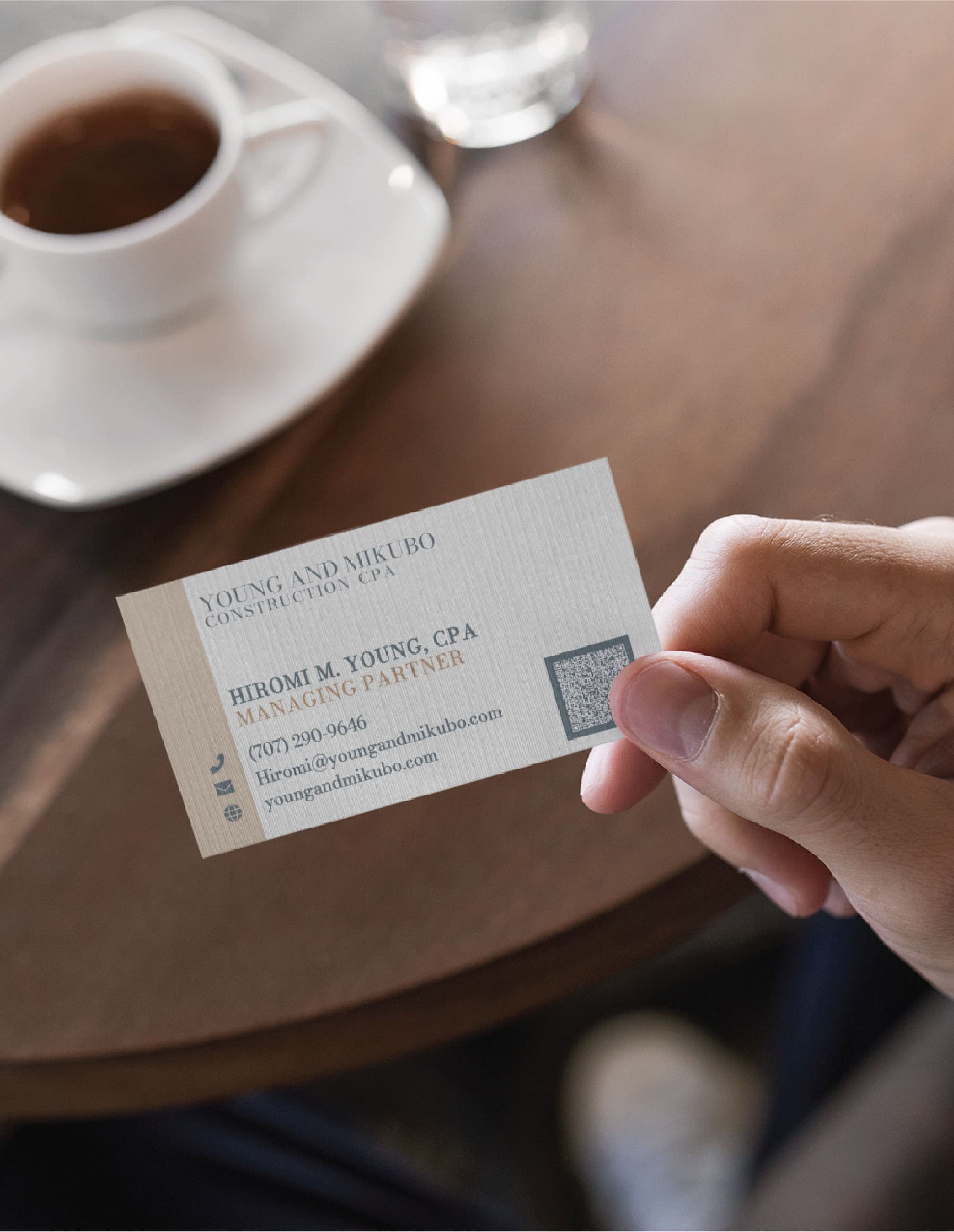 A hand holds a stationery design business card for Hiromi M. Young, Managing Partner at Young and Mikubo Construction CPA, featuring a phone number, email address, website, and a QR code.