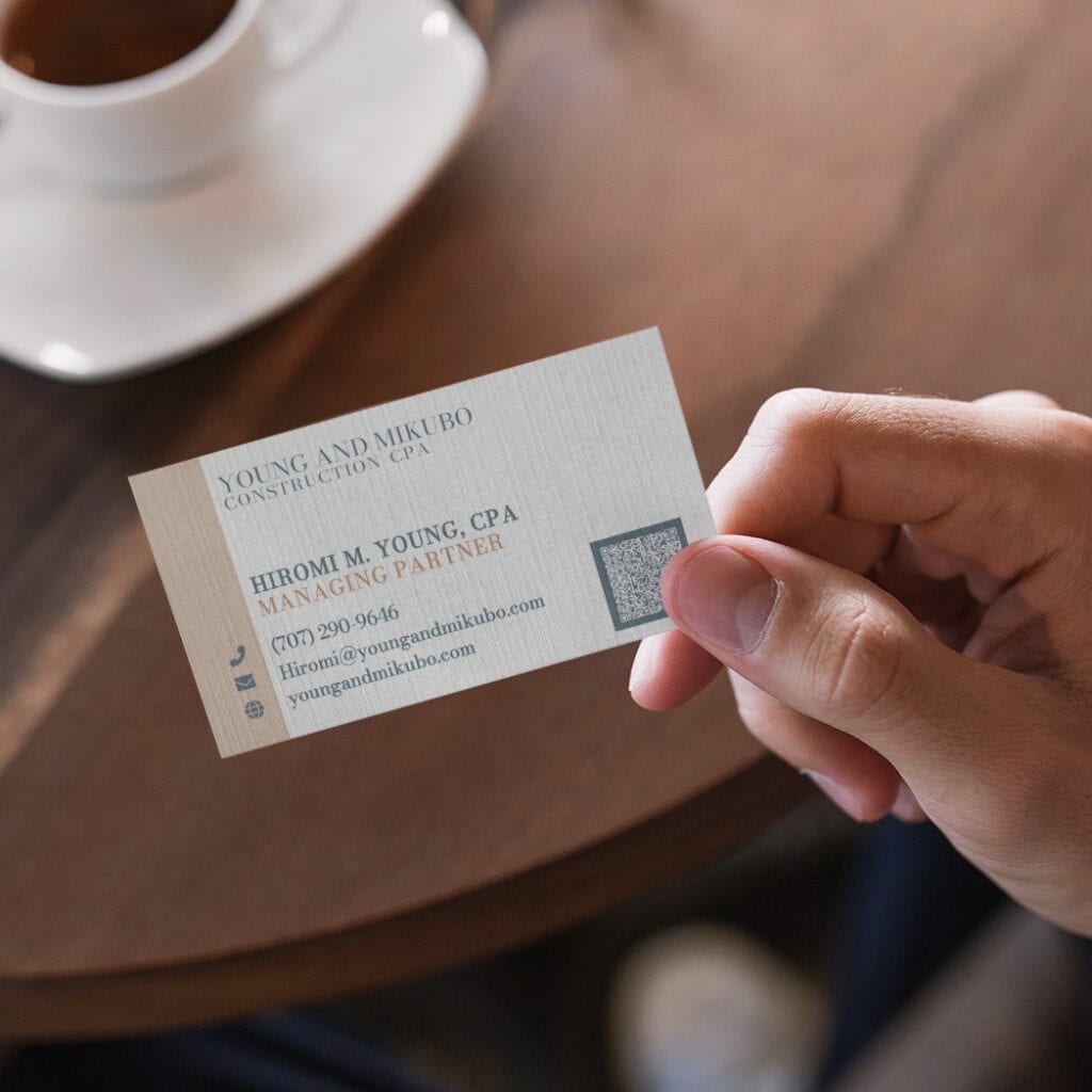 A hand holds a business card for Young and Mikubo Construction CPA, featuring the name Hiromi M. Young, contact information, a QR code, and professional Texas accounting stationery design.