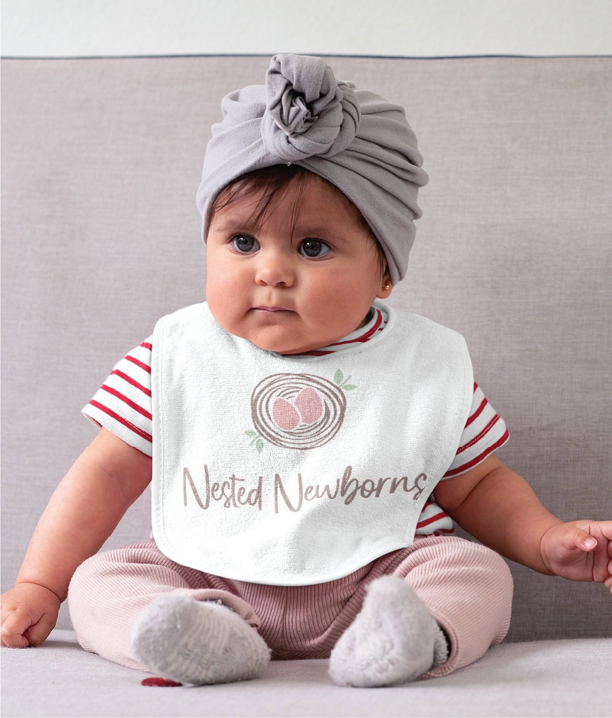 A baby wears a grey turban and a white bib featuring a Nested Newborns logo, an ideal image for a newborn sleep consultant.