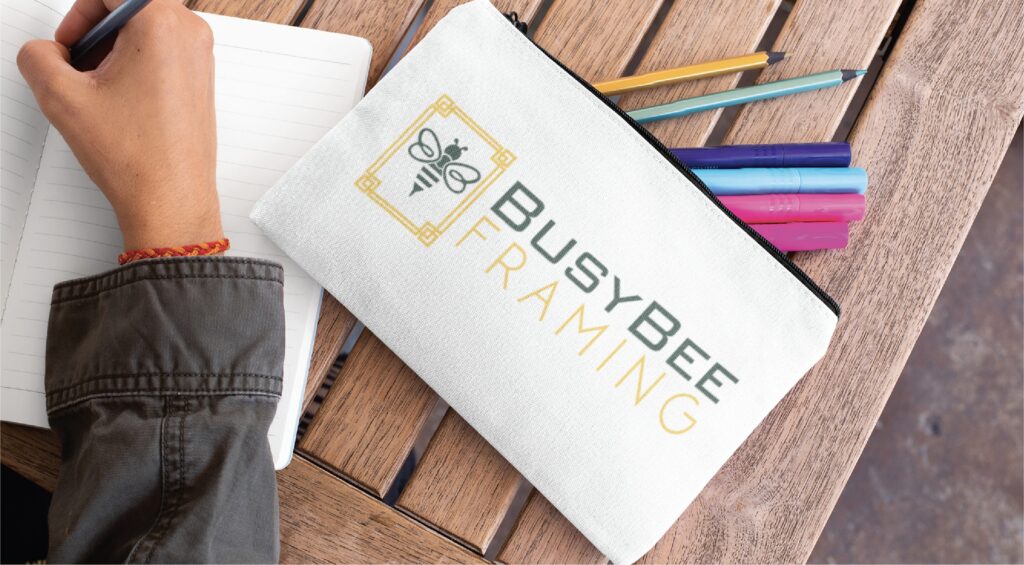 A white zippered pencil pouch featuring professional logo design for Busybee Framing with a bee icon, resting on a wooden table next to a notebook and several colored pens.