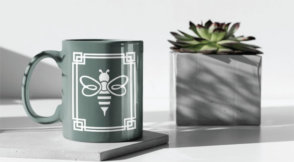 A sage green mug featuring a professional logo design of a white bee inside a square border, sitting on a coaster next to a potted succulent.