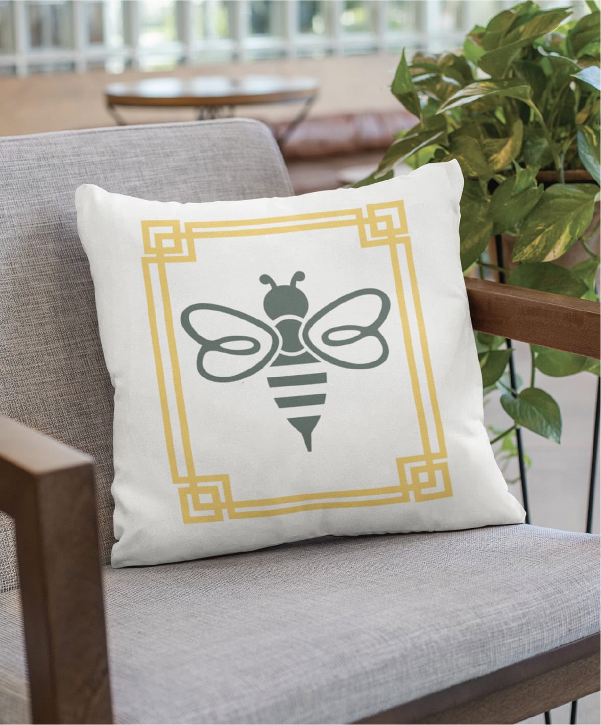 White throw pillow featuring a professional green bee logo design inside a yellow square geometric border, placed on a gray chair.