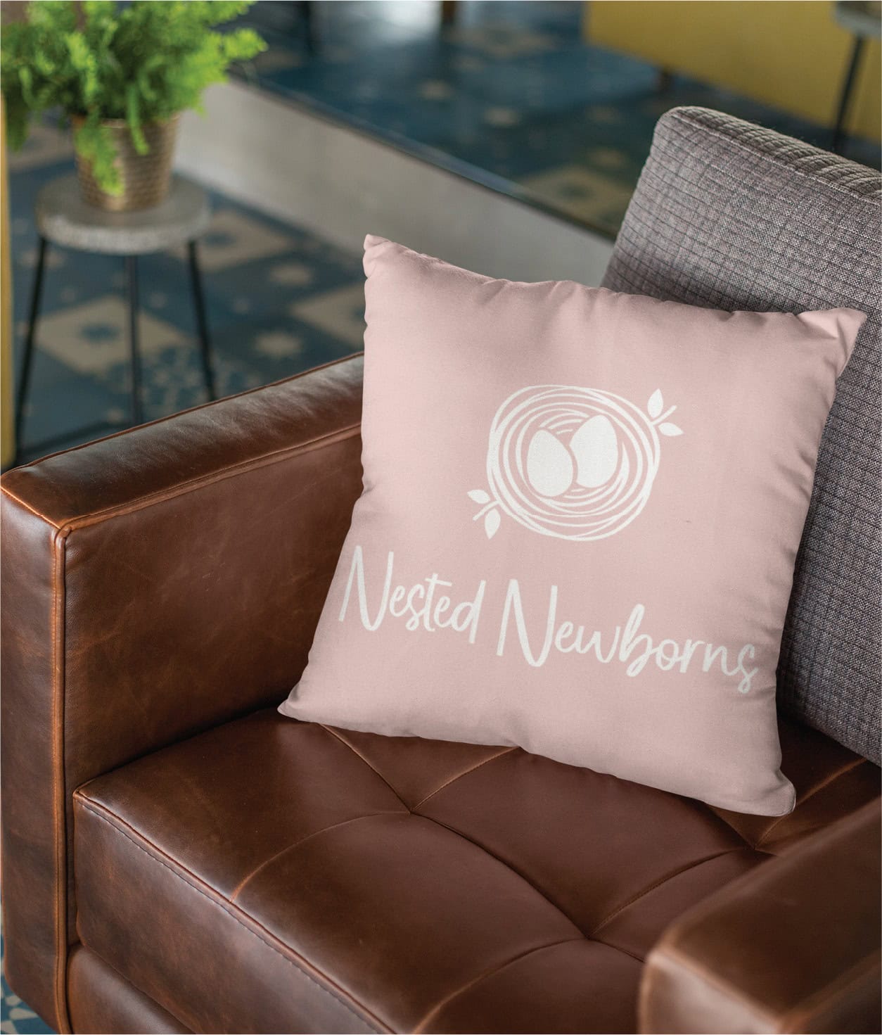 A blush-colored throw pillow featuring a white nest-and-eggs logo for a newborn sleep consultant, resting on a brown leather chair.