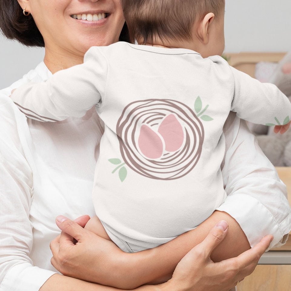 A woman holds a newborn sleep consultant’s baby, who wears a long-sleeved white bodysuit featuring a graphic of two pink eggs in a brown nest.